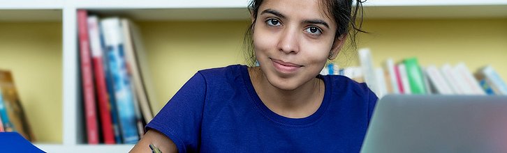 Eine Person vor einem Bücherregal und mit einem Laptop schaut in die Kamera